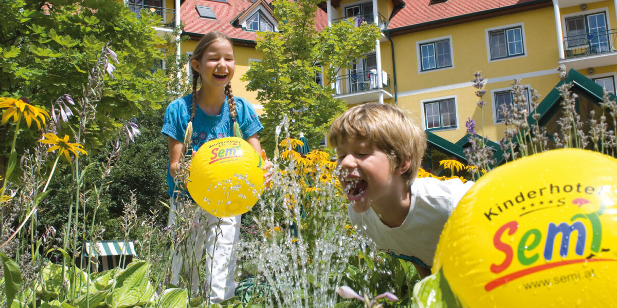 Zwei Kinder spielen mit gelben Strandbällen in der Nähe eines Wasserbrunnens in einem Garten vor einem gelben Gebäude mit rotem Dach und Balkonen an einem sonnigen Tag.