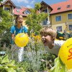 Zwei Kinder spielen mit gelben Strandbällen in der Nähe eines Wasserbrunnens in einem Garten vor einem gelben Gebäude mit rotem Dach und Balkonen an einem sonnigen Tag.