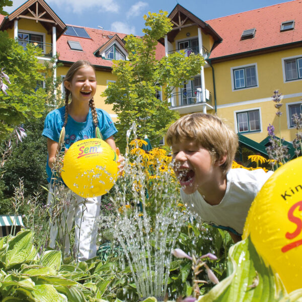 Zwei Kinder spielen mit gelben Strandbällen in der Nähe eines Wasserbrunnens in einem Garten vor einem gelben Gebäude mit rotem Dach und Balkonen an einem sonnigen Tag.