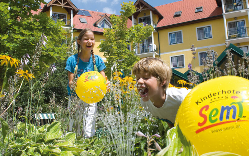 Zwei Kinder spielen mit gelben Strandbällen in der Nähe eines Wasserbrunnens in einem Garten vor einem gelben Gebäude mit rotem Dach und Balkonen an einem sonnigen Tag.