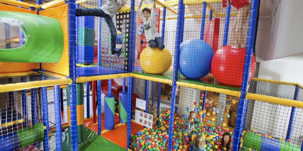 Kinder spielen auf Klettergerüsten und großen bunten Bällen in einem Indoor-Spielplatz mit einer Ballgrube, umgeben von Sicherheitsnetzen.