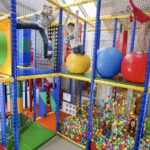 Kinder spielen auf Klettergerüsten und großen bunten Bällen in einem Indoor-Spielplatz mit einer Ballgrube, umgeben von Sicherheitsnetzen.