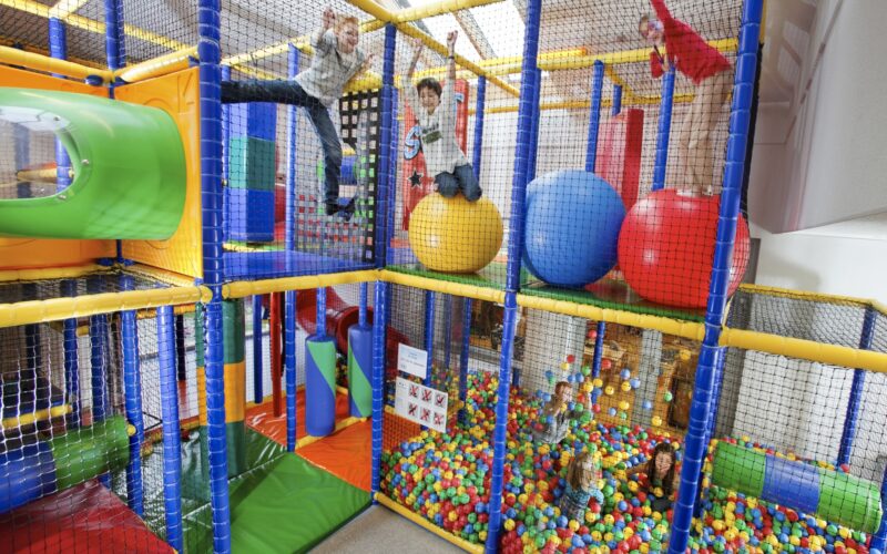 Kinder spielen auf Klettergerüsten und großen bunten Bällen in einem Indoor-Spielplatz mit einer Ballgrube, umgeben von Sicherheitsnetzen.