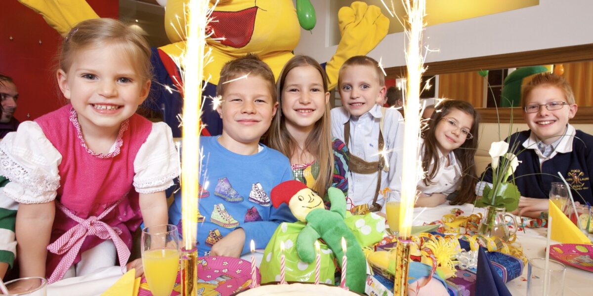 Sechs lächelnde Kinder sitzen an einem geschmückten Tisch mit einer "Kinderhotel Semi"-Torte, umgeben von Partydekoration und Wunderkerzen, und hinter ihnen steht eine Person in einem gelben Maskottchenkostüm.
