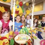 Sechs lächelnde Kinder sitzen an einem geschmückten Tisch mit einer "Kinderhotel Semi"-Torte, umgeben von Partydekoration und Wunderkerzen, und hinter ihnen steht eine Person in einem gelben Maskottchenkostüm.
