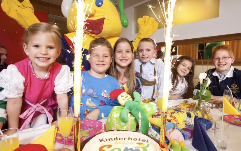 Sechs lächelnde Kinder sitzen an einem geschmückten Tisch mit einer "Kinderhotel Semi"-Torte, umgeben von Partydekoration und Wunderkerzen, und hinter ihnen steht eine Person in einem gelben Maskottchenkostüm.