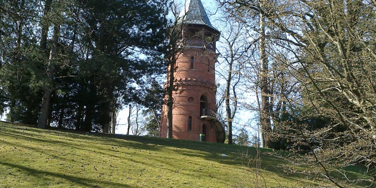 Ein runder, roter Backsteinturm mit kegelförmigem Dach steht auf einem grasbewachsenen Hügel, umgeben von hohen Bäumen unter einem klaren blauen Himmel.