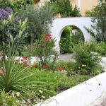 Ein Garten mit verschiedenen blühenden Pflanzen, grünen Sträuchern und einer weißen geschwungenen Mauer mit einem runden Ausschnitt in der Nähe eines gepflasterten Weges.