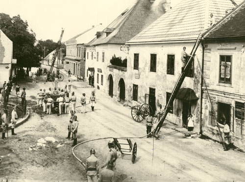 Feuerwehrleute in Uniform bekämpfen mit Leitern und Schläuchen einen Brand in einer von Altbauten gesäumten Straße, während Schaulustige zusehen.