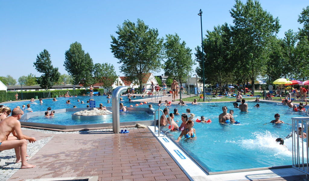 Menschen, die an einem sonnigen Tag in einem öffentlichen Freibad schwimmen und sich entspannen, mit Bäumen und Sitzgelegenheiten am Beckenrand im Hintergrund.