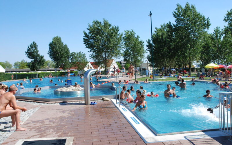 Menschen, die an einem sonnigen Tag in einem öffentlichen Freibad schwimmen und sich entspannen, mit Bäumen und Sitzgelegenheiten am Beckenrand im Hintergrund.