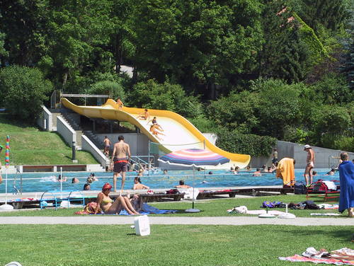 An sonnigen Tagen entspannt man sich in einem Freibad mit Wasserrutsche, umgeben von grünen Bäumen und Gras.
