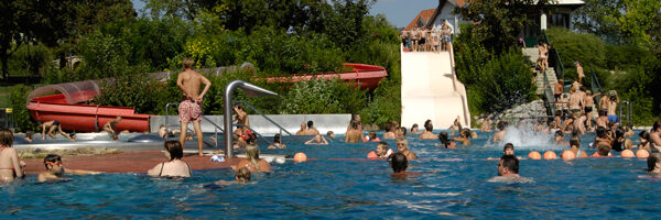 Freibad mit vielen Menschen; einige schwimmen, andere sind in der Nähe der Rutschen und auf dem Pooldeck versammelt. Im Hintergrund sind Bäume und Gebäude zu sehen.