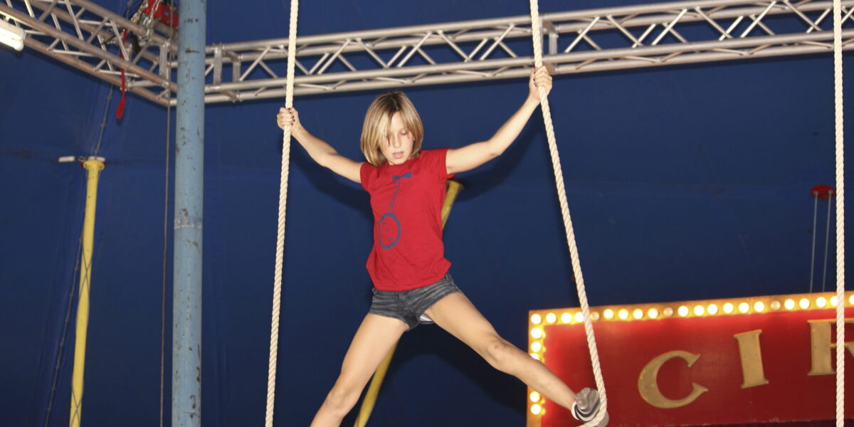 Ein Kind in rotem Hemd und kurzer Hose balanciert auf einem Zirkusseilgerät und hält sich an zwei Seilen unter einem großen blauen Zirkuszelt fest.