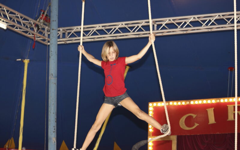 Ein Kind in rotem Hemd und kurzer Hose balanciert auf einem Zirkusseilgerät und hält sich an zwei Seilen unter einem großen blauen Zirkuszelt fest.