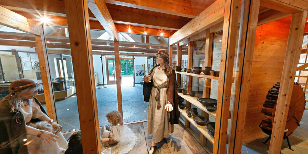 Museumsexponat mit Schaufensterpuppen in historischer Kleidung, Töpferwaren in Regalen und Holzbalken, die die Szene einrahmen.