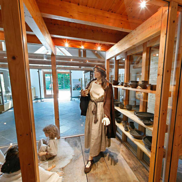 Museumsexponat mit Schaufensterpuppen in historischer Kleidung, Töpferwaren in Regalen und Holzbalken, die die Szene einrahmen.