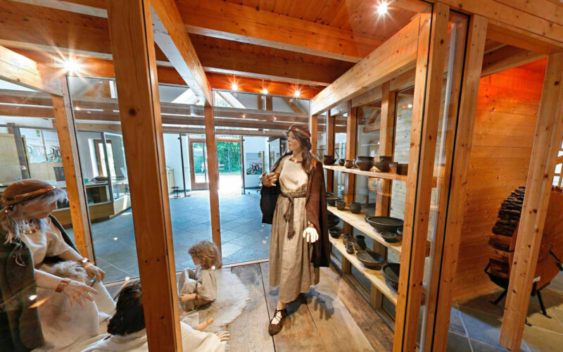 Museumsexponat mit Schaufensterpuppen in historischer Kleidung, Töpferwaren in Regalen und Holzbalken, die die Szene einrahmen.