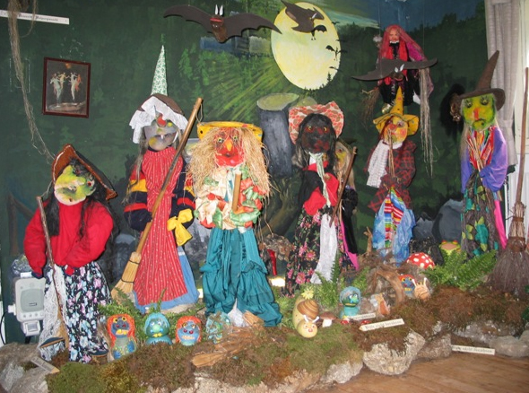 Eine Ausstellung von bunten, menschengroßen Hexenpuppen in Kostümen vor einer Waldkulisse mit Vollmond, Fledermäusen und Waldrequisiten.