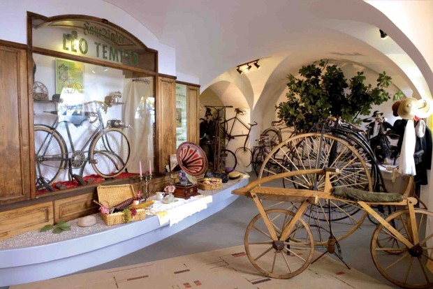 Eine Museumsausstellung mit antiken Fahrrädern, einem hölzernen Dreirad, alten Gegenständen und dekorativen Pflanzen unter Gewölbedecken.