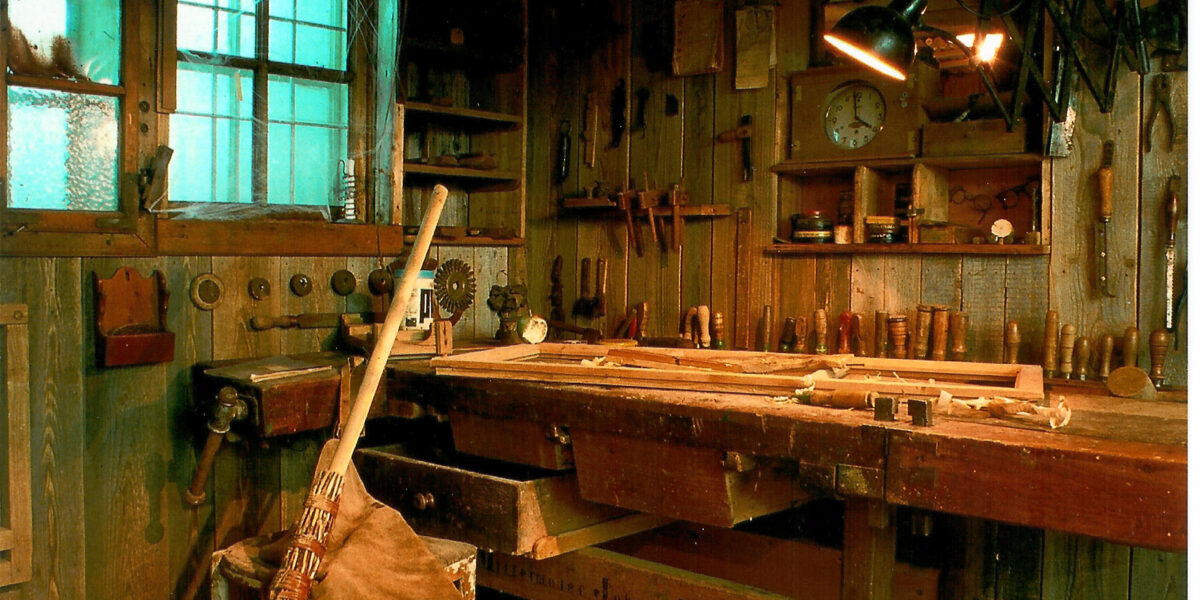 Eine rustikale Holzwerkstatt mit an den Wänden hängenden Werkzeugen, einer mit Werkzeugen bedeckten Werkbank, einem Besen, einer Lederschürze und auf dem Holzboden verstreuten Holzspänen.