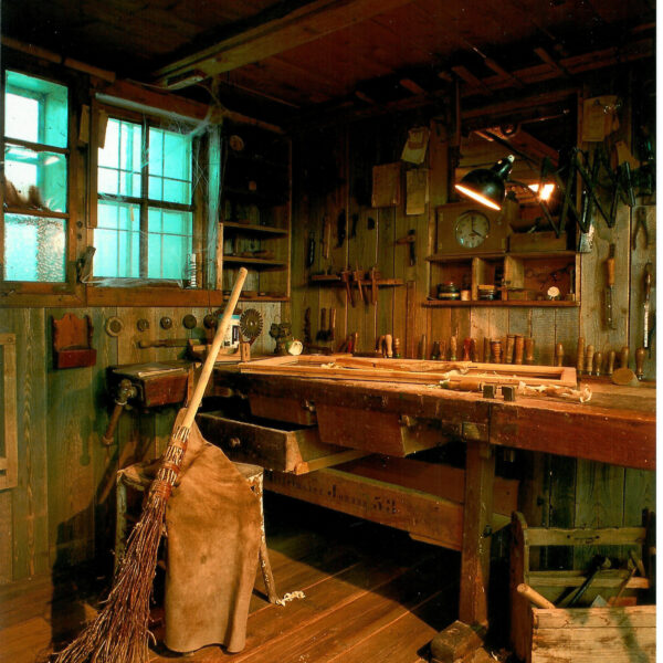 Eine rustikale Holzwerkstatt mit an den Wänden hängenden Werkzeugen, einer mit Werkzeugen bedeckten Werkbank, einem Besen, einer Lederschürze und auf dem Holzboden verstreuten Holzspänen.