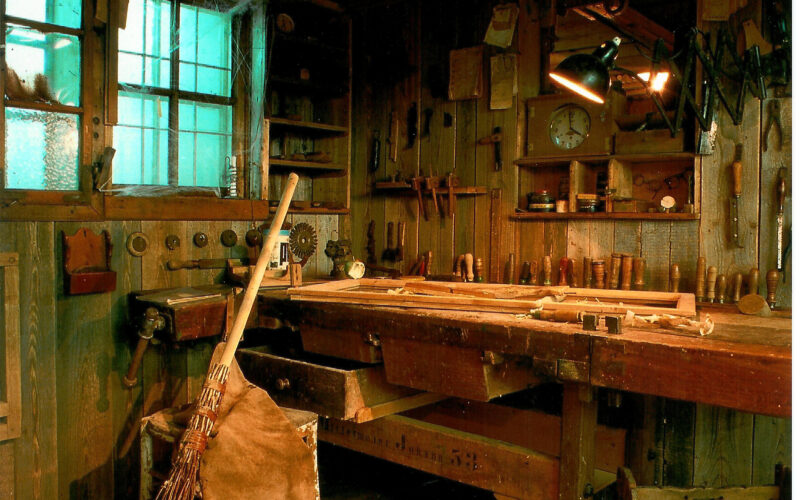 Eine rustikale Holzwerkstatt mit an den Wänden hängenden Werkzeugen, einer mit Werkzeugen bedeckten Werkbank, einem Besen, einer Lederschürze und auf dem Holzboden verstreuten Holzspänen.