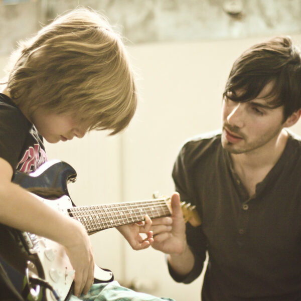 Ein Erwachsener bringt einem Kind das Spielen einer E-Gitarre bei, indem er in einem gut beleuchteten Raum die Hand des Kindes auf dem Griffbrett führt.