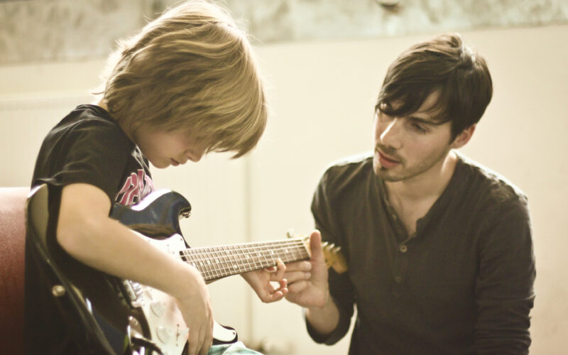 Ein Erwachsener bringt einem Kind das Spielen einer E-Gitarre bei, indem er in einem gut beleuchteten Raum die Hand des Kindes auf dem Griffbrett führt.