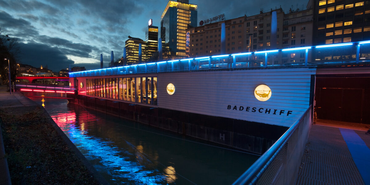 Ein modernes schwimmendes Bauwerk mit der Aufschrift "Badeschiff" und blauer Akzentbeleuchtung liegt in der Abenddämmerung in einem Fluss neben den Gebäuden der Stadt vertäut.