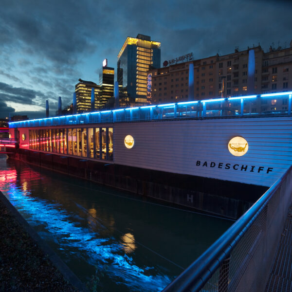 Ein modernes schwimmendes Bauwerk mit der Aufschrift "Badeschiff" und blauer Akzentbeleuchtung liegt in der Abenddämmerung in einem Fluss neben den Gebäuden der Stadt vertäut.