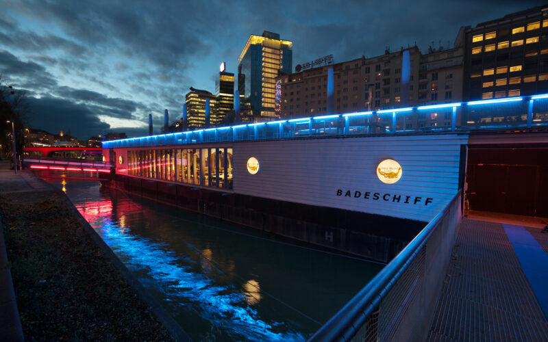 Ein modernes schwimmendes Bauwerk mit der Aufschrift "Badeschiff" und blauer Akzentbeleuchtung liegt in der Abenddämmerung in einem Fluss neben den Gebäuden der Stadt vertäut.