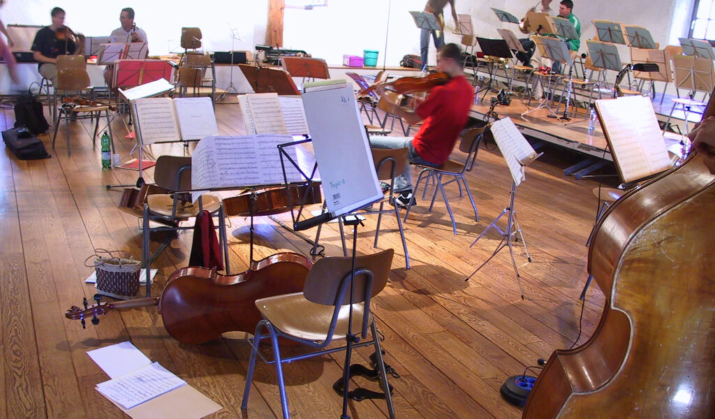Mehrere Musiker in einem Proberaum mit verstreuten Stühlen, Notenständern, Notenblättern und Streichinstrumenten, darunter Bässe und Geigen.
