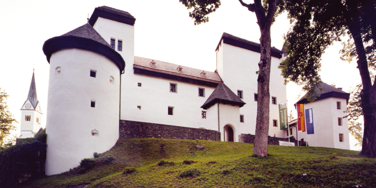 Ein weißes Schloss im mittelalterlichen Stil mit runden Türmen und einem abfallenden Grashügel davor, der teilweise von großen Bäumen beschattet wird.