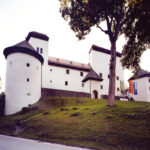 Ein weißes Schloss im mittelalterlichen Stil mit runden Türmen und einem abfallenden Grashügel davor, der teilweise von großen Bäumen beschattet wird.