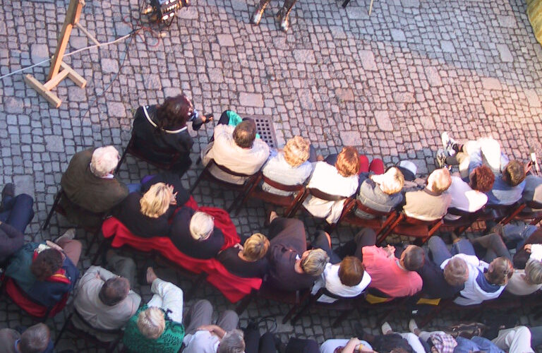 Ein Künstler steht vor einem sitzenden Publikum, das in Reihen auf einem kopfsteingepflasterten Innenhof angeordnet ist, von oben gesehen.