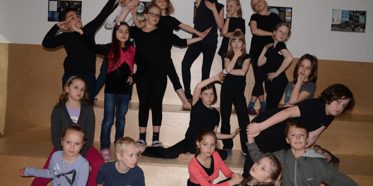 Eine Gruppe von Kindern posiert auf einer hölzernen Treppe. Die meisten von ihnen sind schwarz gekleidet und nehmen dramatische oder spielerische Posen für die Kamera ein.