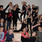 Eine Gruppe von Kindern posiert auf einer hölzernen Treppe. Die meisten von ihnen sind schwarz gekleidet und nehmen dramatische oder spielerische Posen für die Kamera ein.