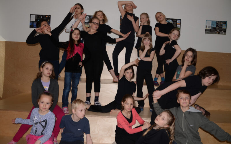 Eine Gruppe von Kindern posiert auf einer hölzernen Treppe. Die meisten von ihnen sind schwarz gekleidet und nehmen dramatische oder spielerische Posen für die Kamera ein.