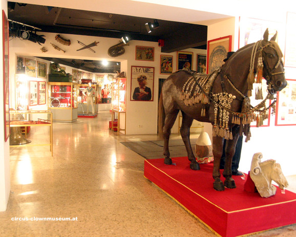 Museumsausstellung mit einer lebensgroßen Pferdestatue, die mit dekorativen Geschirren geschmückt ist, umgeben von Zirkusandenken und Vitrinen.