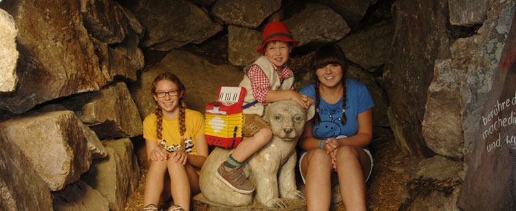 Drei Kinder sitzen in einer Steinhöhle; eines sitzt auf einer weißen Bärenstatue, während die beiden anderen an den Seiten sitzen und in die Kamera lächeln.