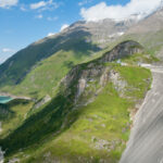 Ein großer geschwungener Staudamm staut türkisfarbenes Wasser in einem Bergtal, umgeben von steilen, grünen Hängen und teilweise schneebedeckten Gipfeln unter blauem Himmel.