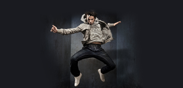 Eine Person in legerer Kleidung und weißen Turnschuhen befindet sich in der Luft und springt energisch vor einem dunklen, minimalistischen Hintergrund.