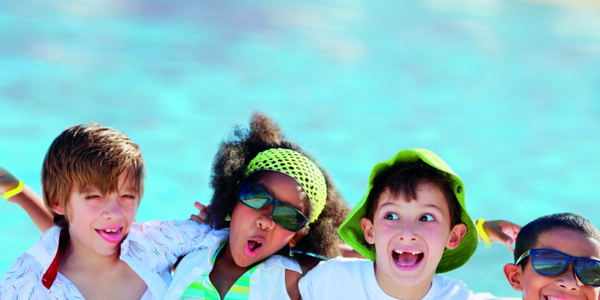Vier Kinder in Sommerkleidung und mit Sonnenbrille stehen lächelnd und grinsend vor einem Schwimmbad.