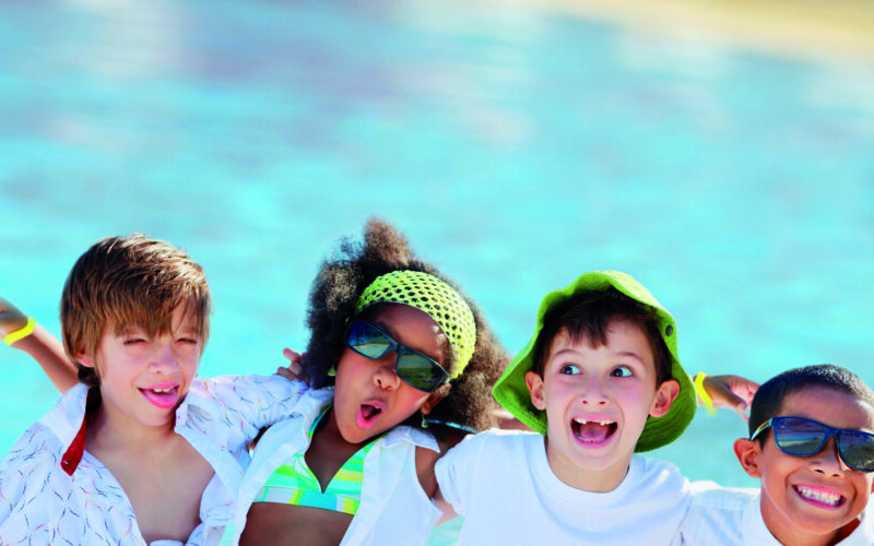 Vier Kinder in Sommerkleidung und mit Sonnenbrille stehen lächelnd und grinsend vor einem Schwimmbad.