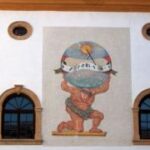 Ein Wandgemälde von Atlas, der kniend eine Weltkugel mit einer Sonnenuhr in der Hand hält, befindet sich zwischen zwei Bogenfenstern an einer weißen Gebäudefassade.