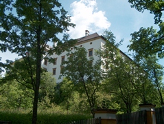 Ein weißes, mehrstöckiges Gebäude mit mehreren Fenstern wird teilweise von hohen grünen Bäumen unter einem blauen Himmel mit einigen Wolken verdeckt.