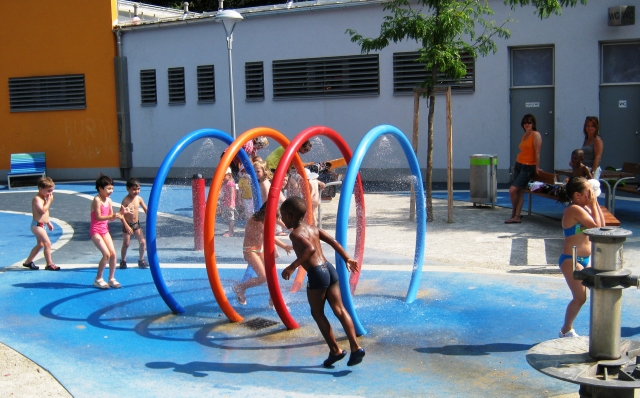 Kinder in Badeanzügen spielen an einem sonnigen Tag in einem Wasserpark und rennen durch bunte, runde Sprinkler, während Erwachsene in der Nähe zuschauen.