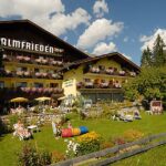 Ein großes Alpenhotel mit blumengeschmückten Balkonen, einem grünen Rasen mit Gartenmöbeln und Menschen, die sich an einem sonnigen Tag im Freien entspannen.