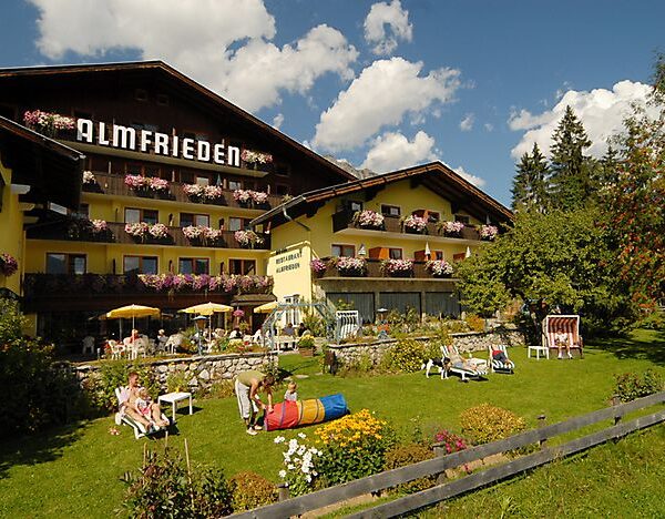 Ein großes Alpenhotel mit blumengeschmückten Balkonen, einem grünen Rasen mit Gartenmöbeln und Menschen, die sich an einem sonnigen Tag im Freien entspannen.